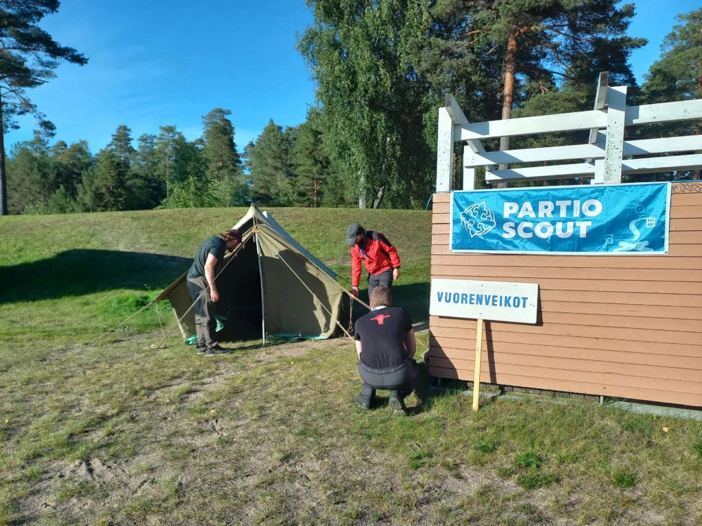 Kolme miestä pystyttämässä telttaa nurmikolle uimakopin viereen