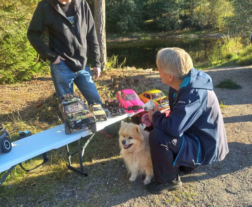 Mies, jonka edessä on penkillä RC-autoja. penkin edessä on vaalea koira, ja toinen mies
